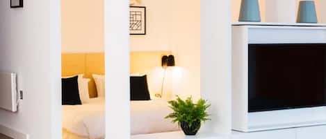 Una habitación de hotel moderna con una cama, una lámpara de noche, una planta en maceta y un televisor de pantalla plana montado sobre un mueble blanco.