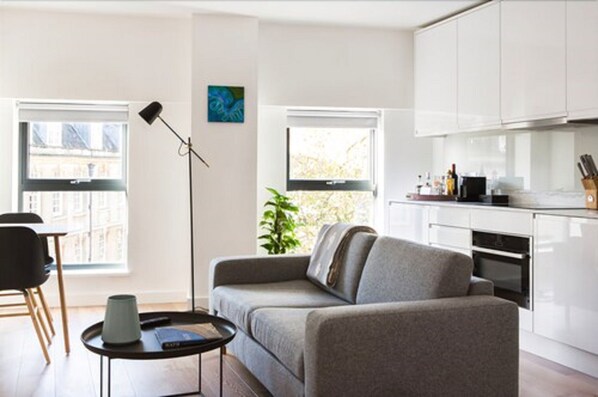 A modern living room with a sofa, a coffee table, and a floor lamp. There is a kitchen area with white cabinets and a window. - Hiding Space - Westgate Apartments (Bath)