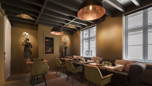 Een gezellige eethoek met een houten tafel, groene stoelen en een bank. Er staan potplanten en is een open haard.