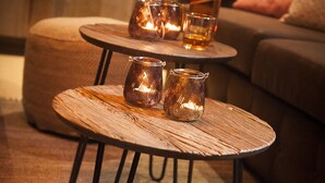 Een gezellige zithoek met twee ronde houten tafels, elk met metalen poten. Op de tafels staan twee kandelaars van glas, en op de bovenste tafel staat een fles drank. De kamer heeft een warme sfeer, met een houten wand op de achtergrond en een bruine bank met kussens.