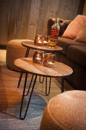 Un espace de vie chaleureux avec deux petites tables rondes en bois, chacune dotée de pieds en métal. Sur les tables, on trouve deux photophores en verre, et une bouteille de spiritueux est posée sur la table du haut. La pièce dégage une atmosphère accueillante, avec un mur en bois en arrière-plan et un canapé marron agrémenté de coussins.