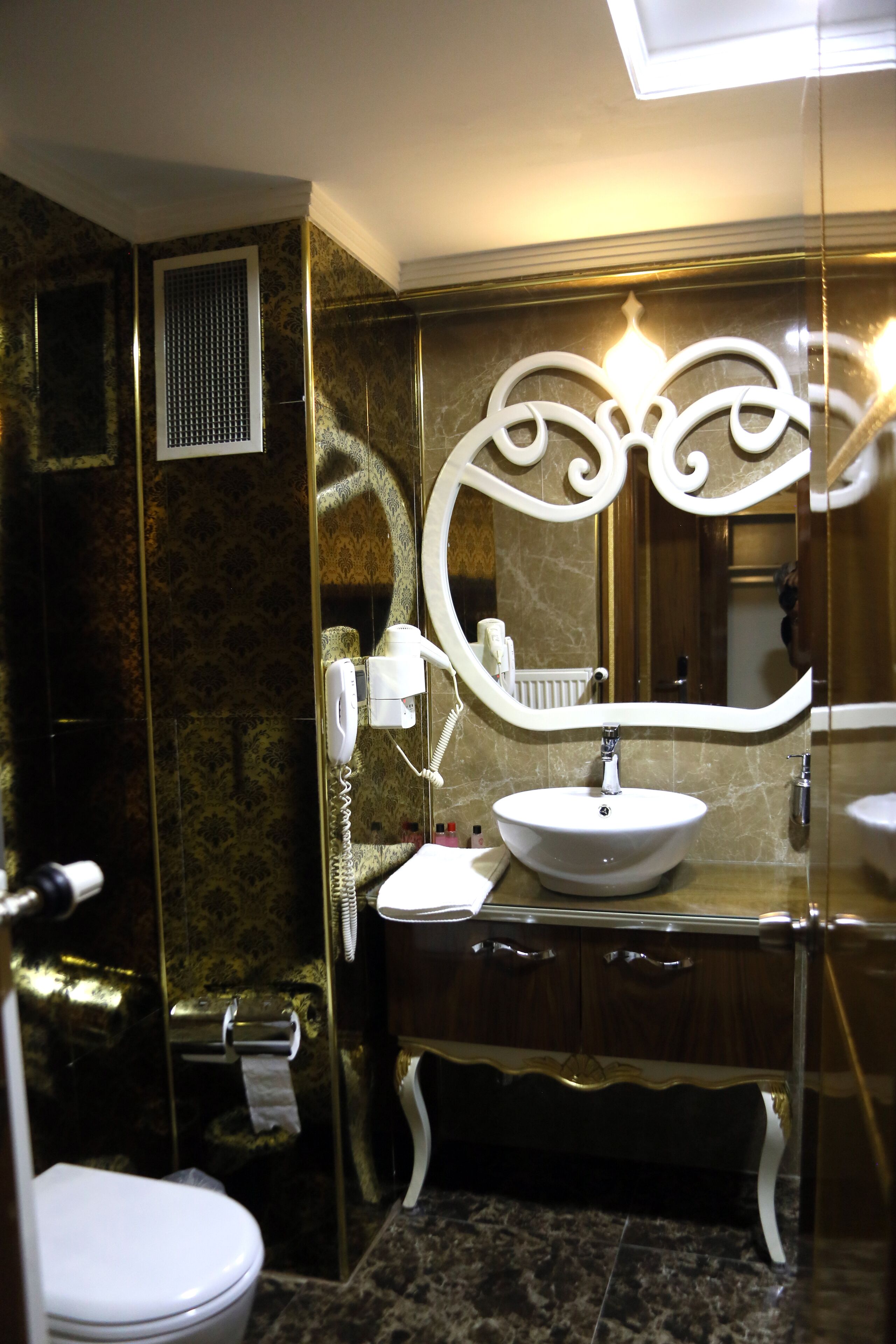 Un baño con pared de mármol, un lavabo blanco, un mueble de tocador con cajones, un espejo y un inodoro.