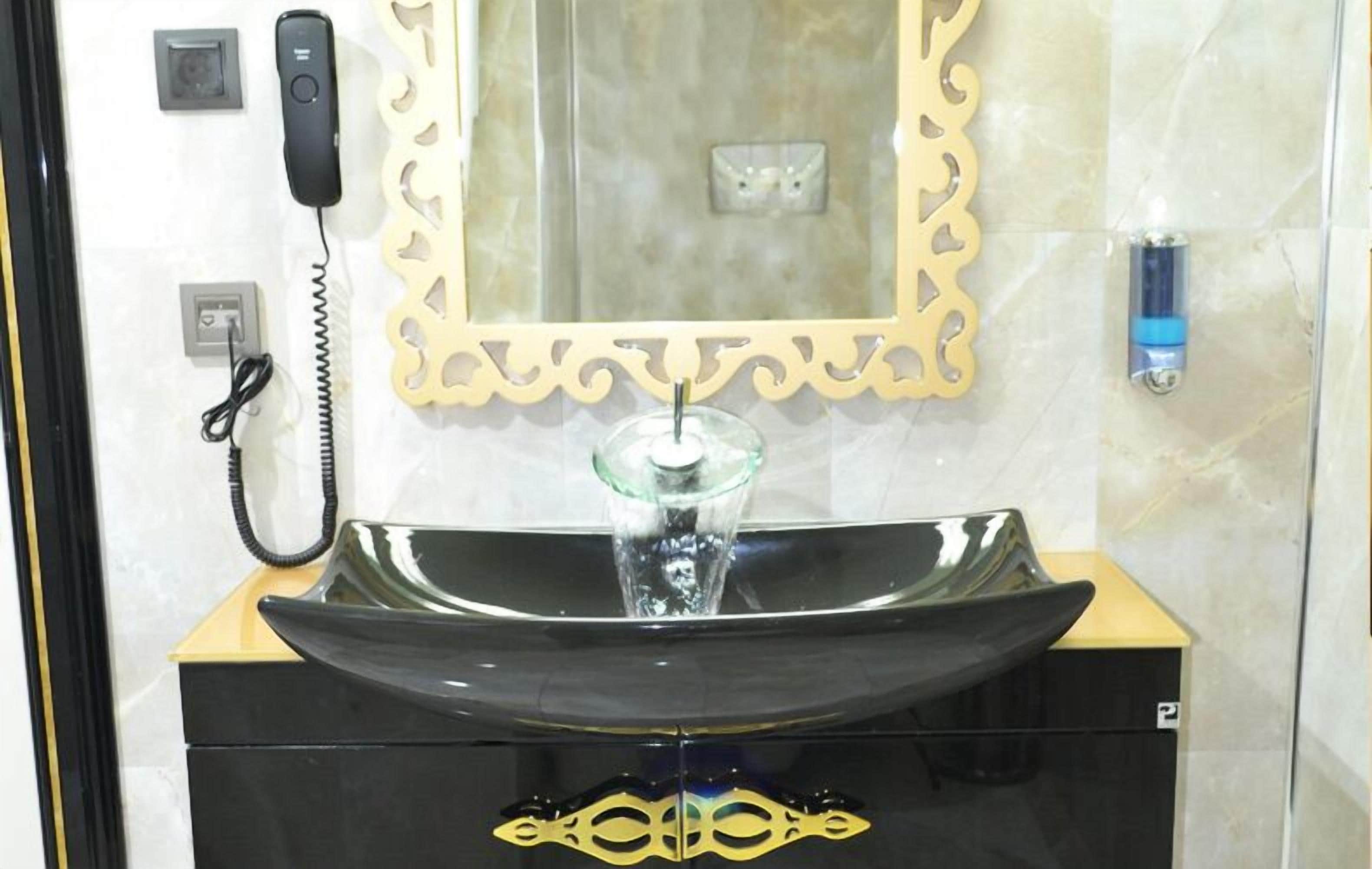 Uma casa de banho com toucador preto, lavatório de vidro, espelho com moldura decorativa, telefone e dispensador de sabonete.