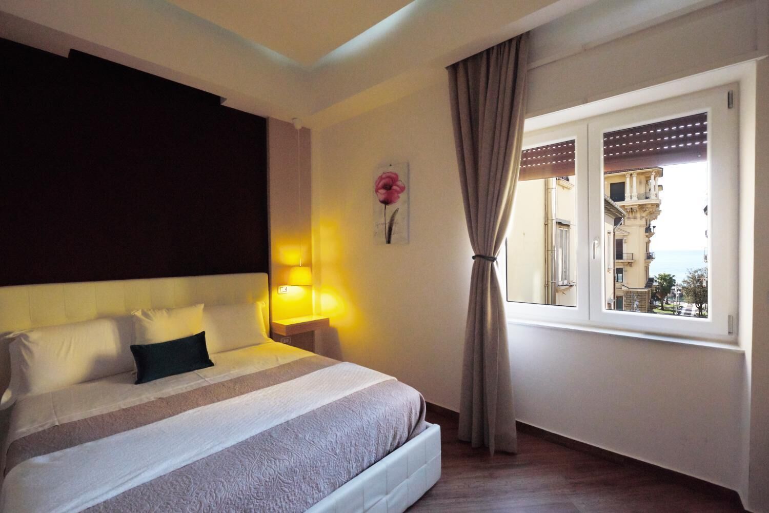 A hotel room with a bed, a nightstand, a lamp, a window with curtains, and a view of buildings.