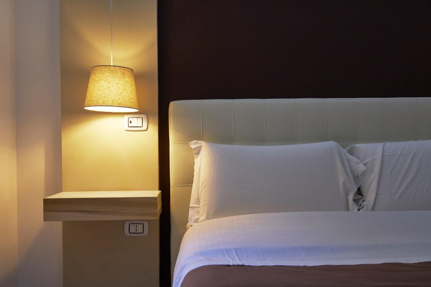 A neatly made bed with white linens and pillows, a wall-mounted lamp, and a power outlet on the nightstand.