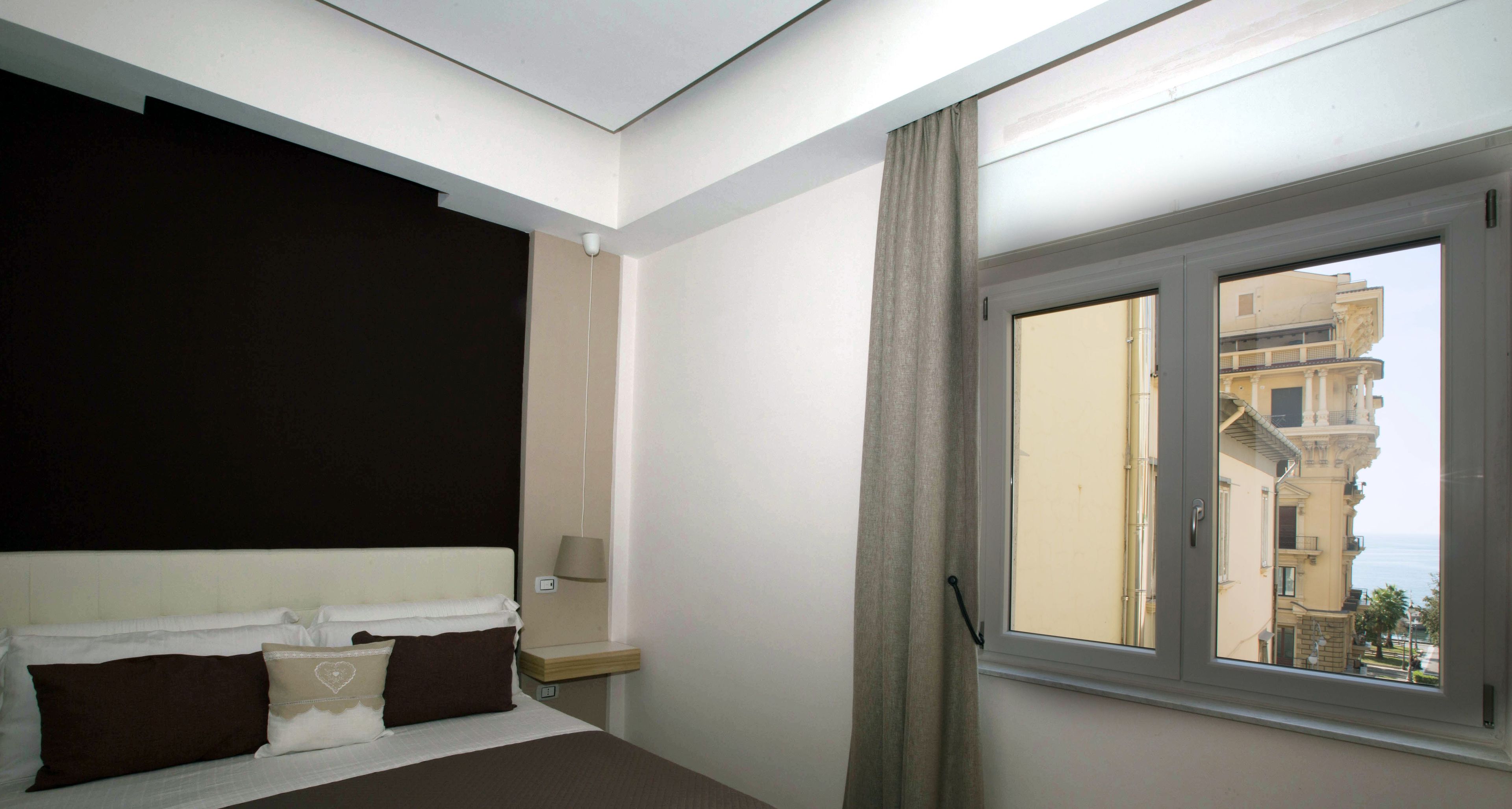 Ein Schlafzimmer mit Bett, Nachttisch, Lampe und Fenster mit Blick auf Gebäude.