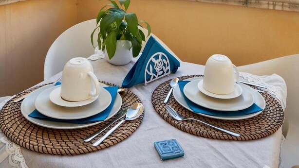 A balcony with a table set for two, featuring white plates, napkins, and cutlery. A potted plant and a blue napkin with a logo are on the table. - 360 Hospitality (Salerno)