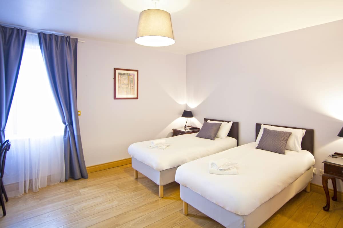 A hotel room with two single beds, a wooden floor, a window with blue curtains, a wall-mounted picture, and a ceiling lamp.