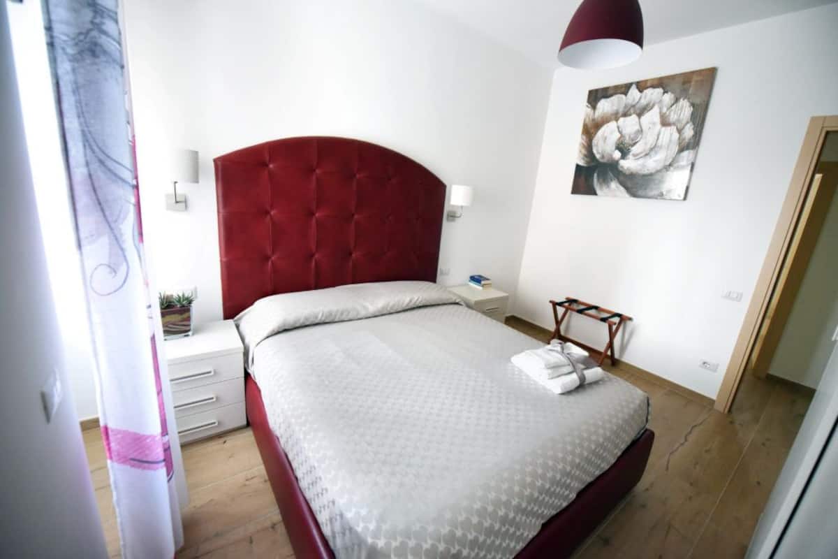Une chambre à coucher avec une tête de lit rouge, un couvre-lit blanc, une table de chevet avec une lampe et une œuvre d’art accrochée au mur.