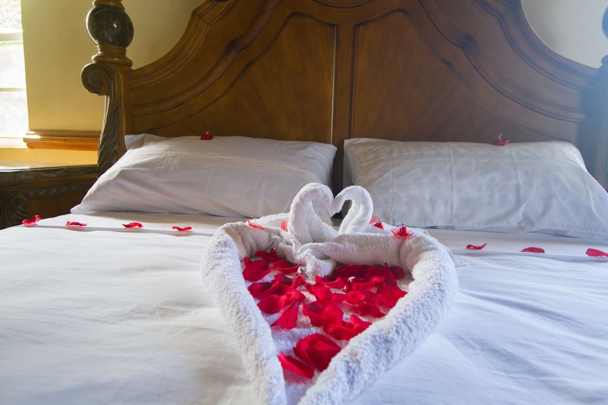 A bed with white linens and a heart-shaped towel with rose petals.