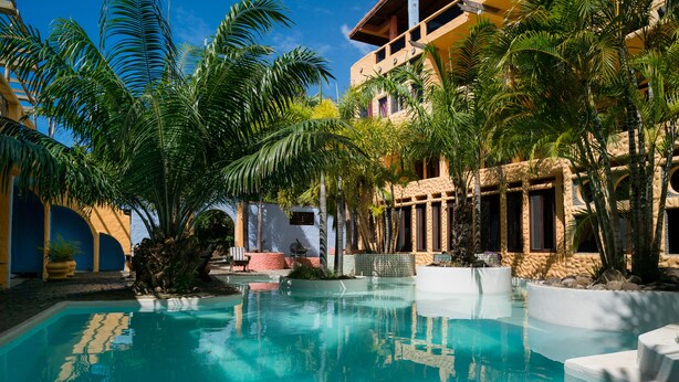 Zona de piscina con agua cristalina, rodeada de palmeras y un edificio de fachada amarilla.