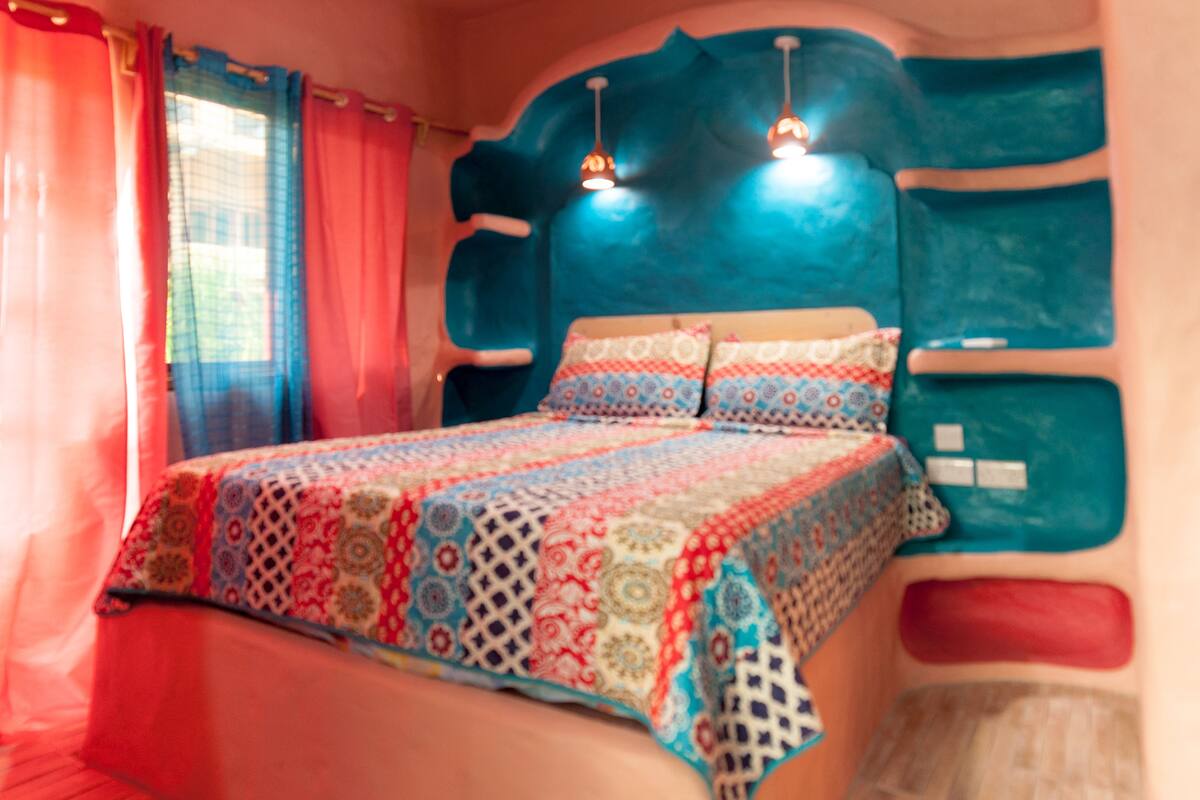 Ein Bett mit farbig gemustertem Überwurf, einem Kopfende mit markantem Design und zwei Pendelleuchten.