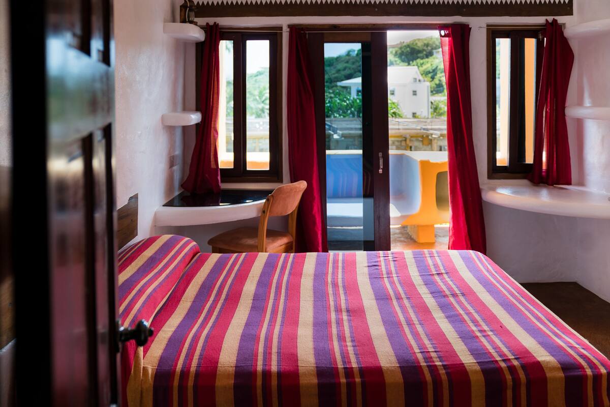 Ein Hotelzimmer mit Bett, Stuhl, kleinem Tisch und Blick ins Freie.