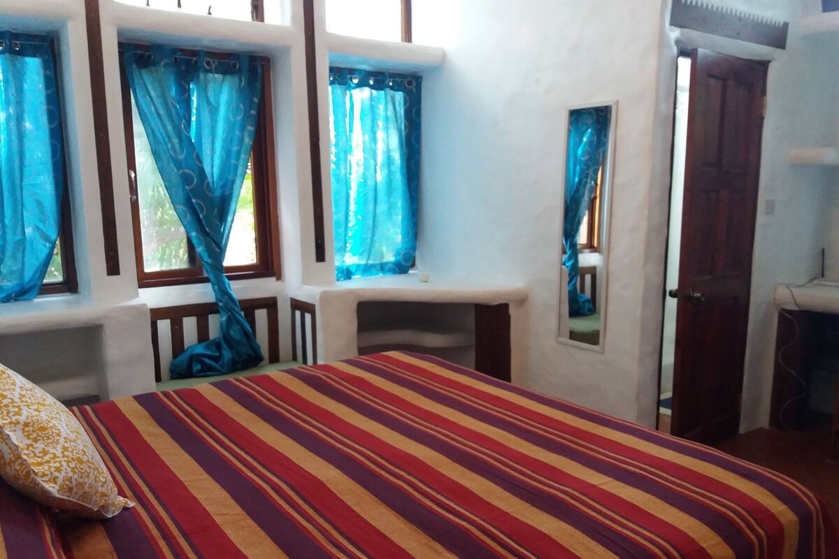 Ein Schlafzimmer mit gestreifter Bettdecke, einem Holzkopfende, einem kleinen Schreibtisch und einem Fenster mit blauen Vorhängen.