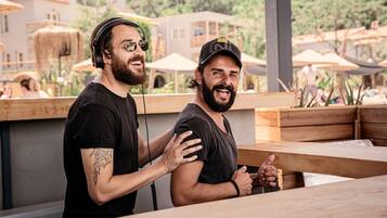 Dos hombres sentados en una mesa de madera con vista a edificios y sombrillas al fondo.