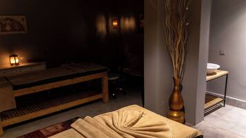 Una habitación tenuemente iluminada con una camilla de masajes, un banco de madera, un jarrón con ramas y un escritorio pequeño con una lámpara.