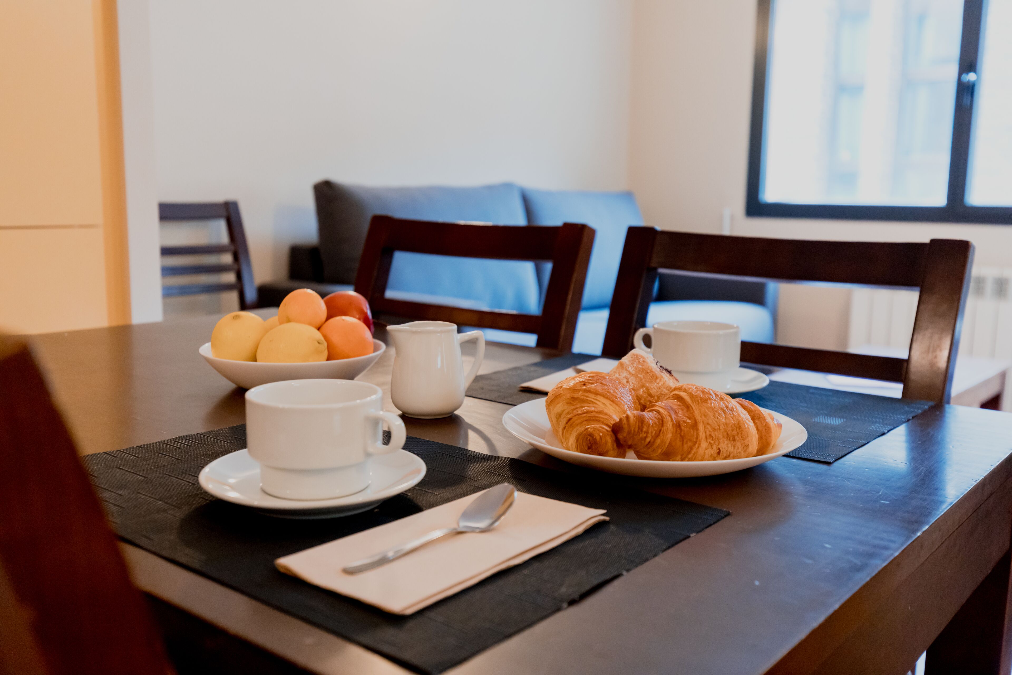 Una mesa de desayuno con cruasanes, un bol de fruta, una taza de café y una tetera.