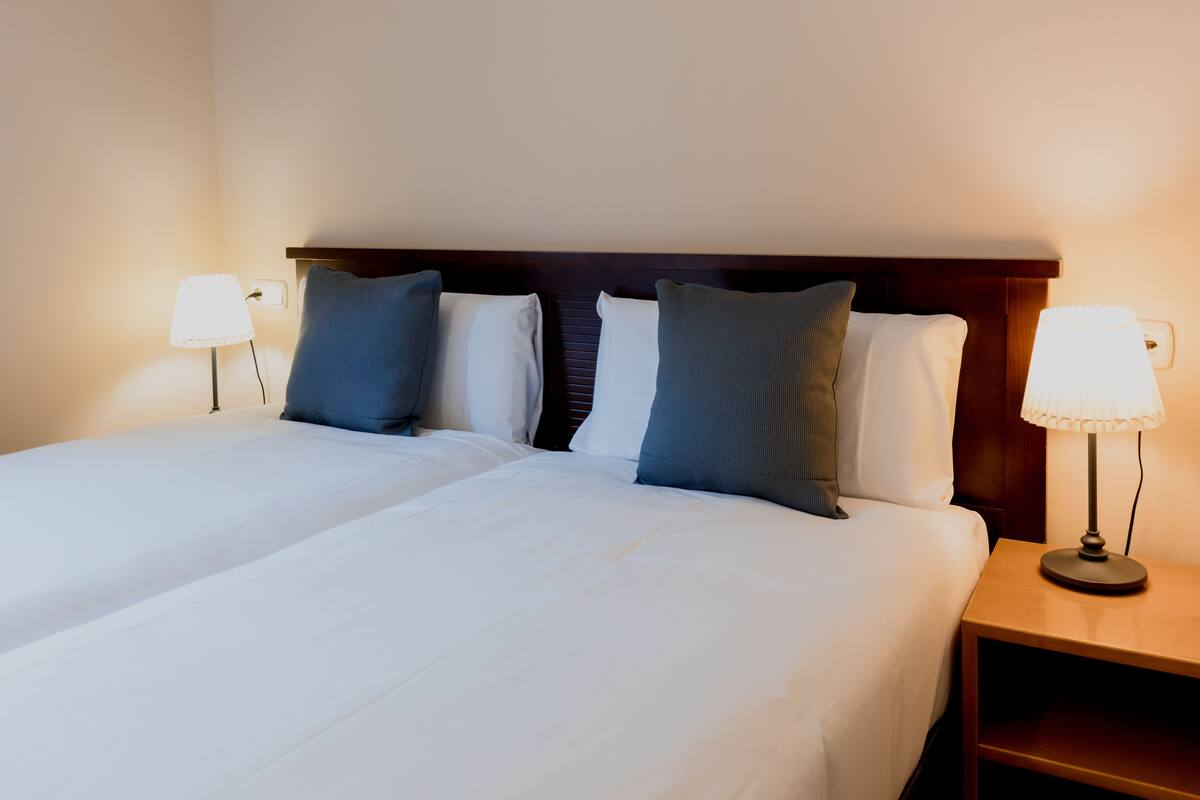 A double bed with white linens, two bedside lamps, and a wooden nightstand.