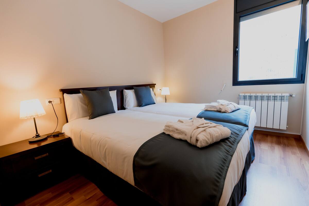 A hotel room with two beds, a nightstand with a lamp, a window with a view, and a radiator.