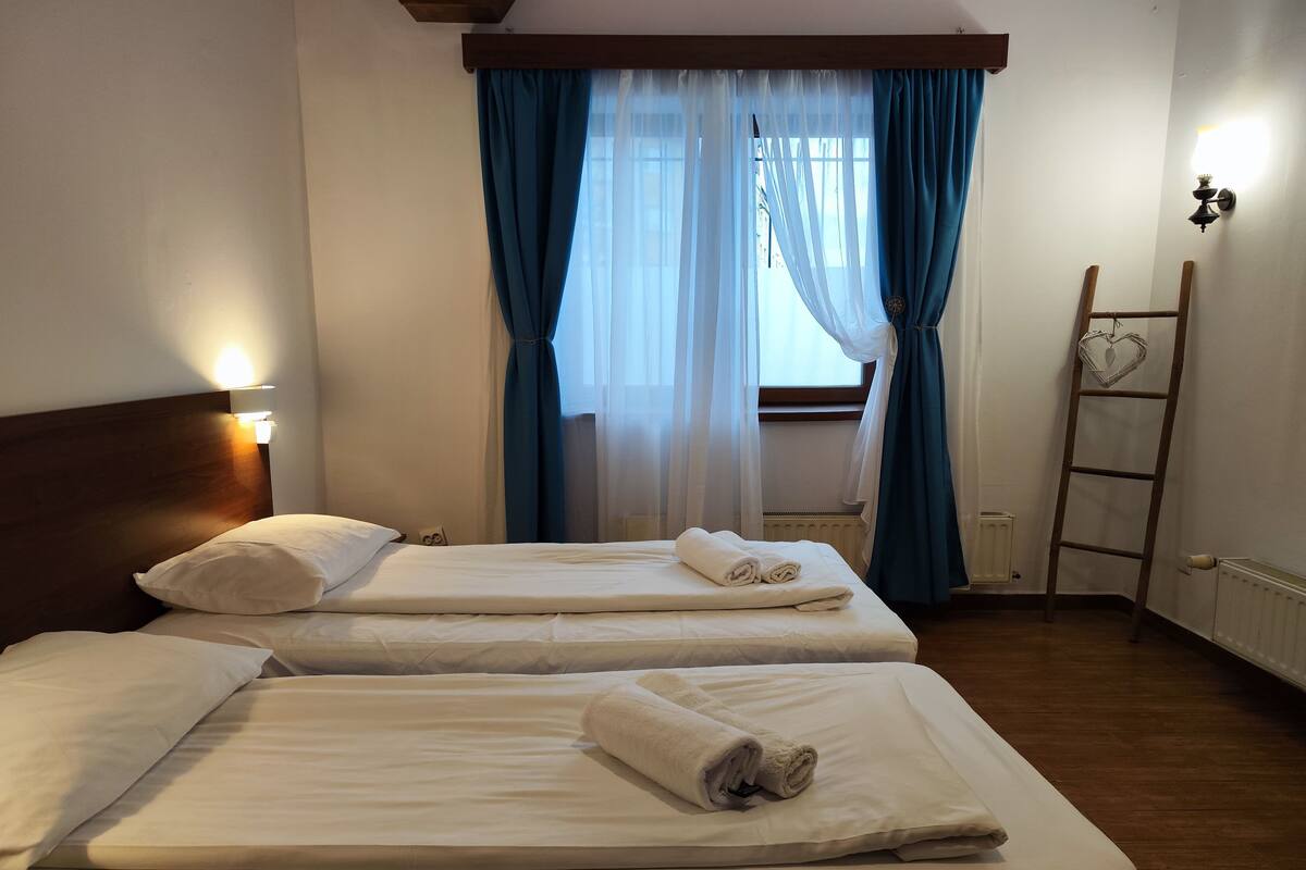 A hotel room with two beds, a wooden headboard, a window with blue curtains, a radiator, and a ladder.