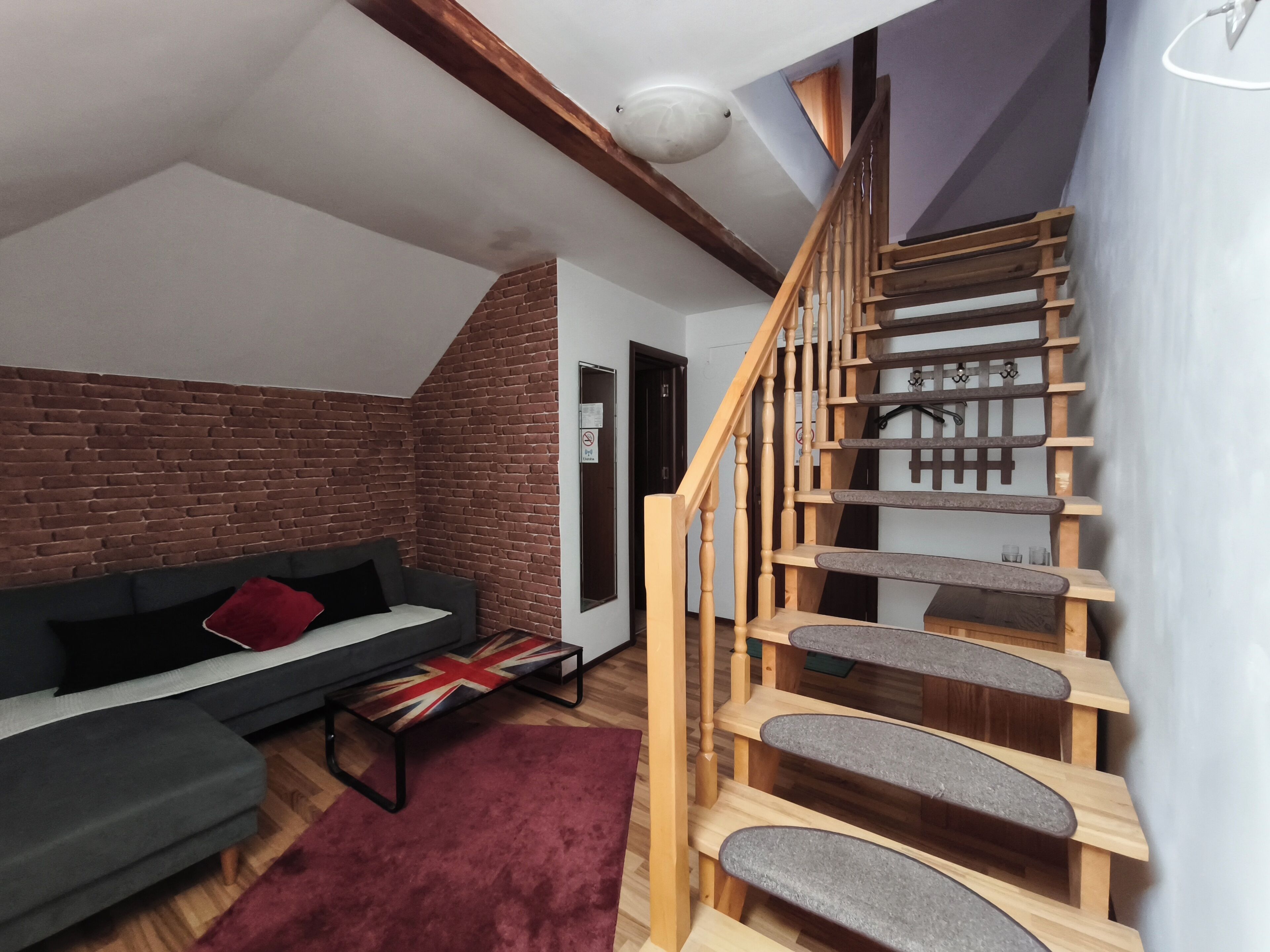 A loft-style living room with a brick wall, a wooden staircase, a grey sofa, and a red rug.