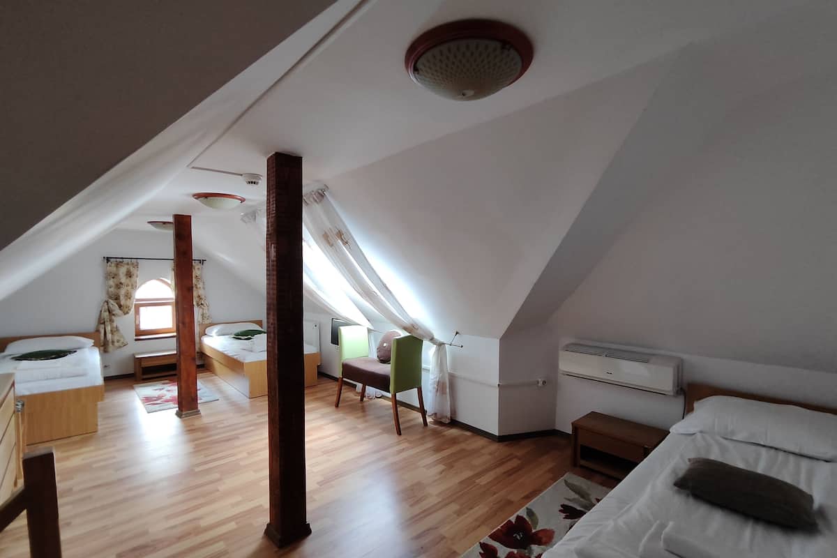 A room with a sloped ceiling, wooden flooring, a bed with pillows, a chair, a small table, and a window with curtains.