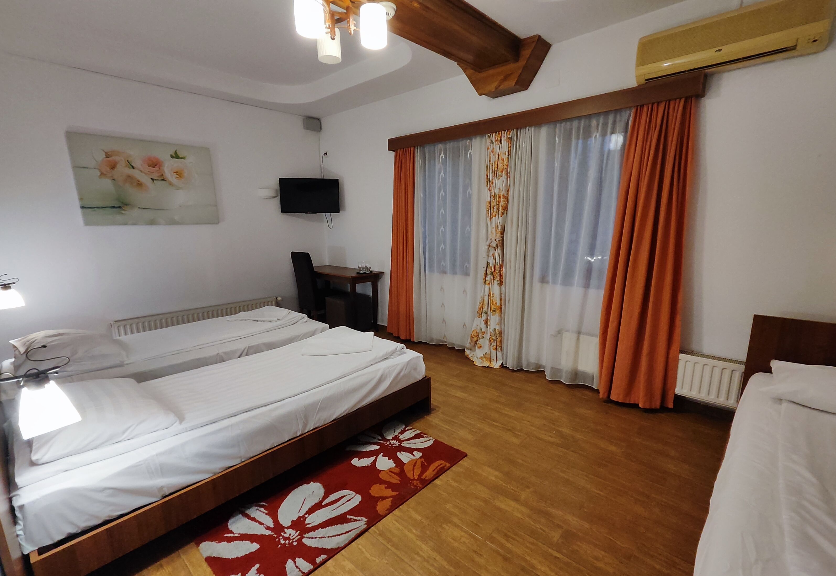 A double bed with a wooden headboard, a bedside table, a red and white floral rug, a wall-mounted TV, and a window with orange curtains.