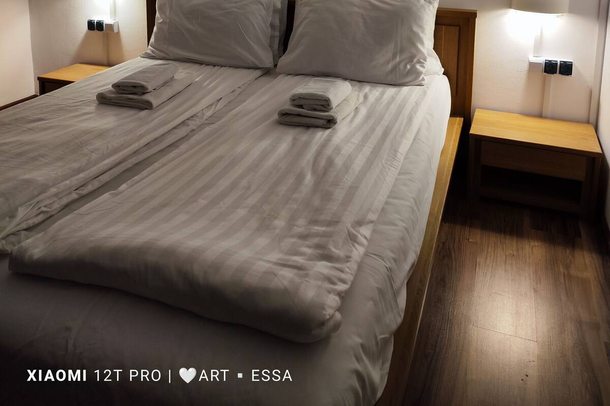 A neatly made bed in a room with a sloped ceiling, two bedside lamps, and a small wooden nightstand.
