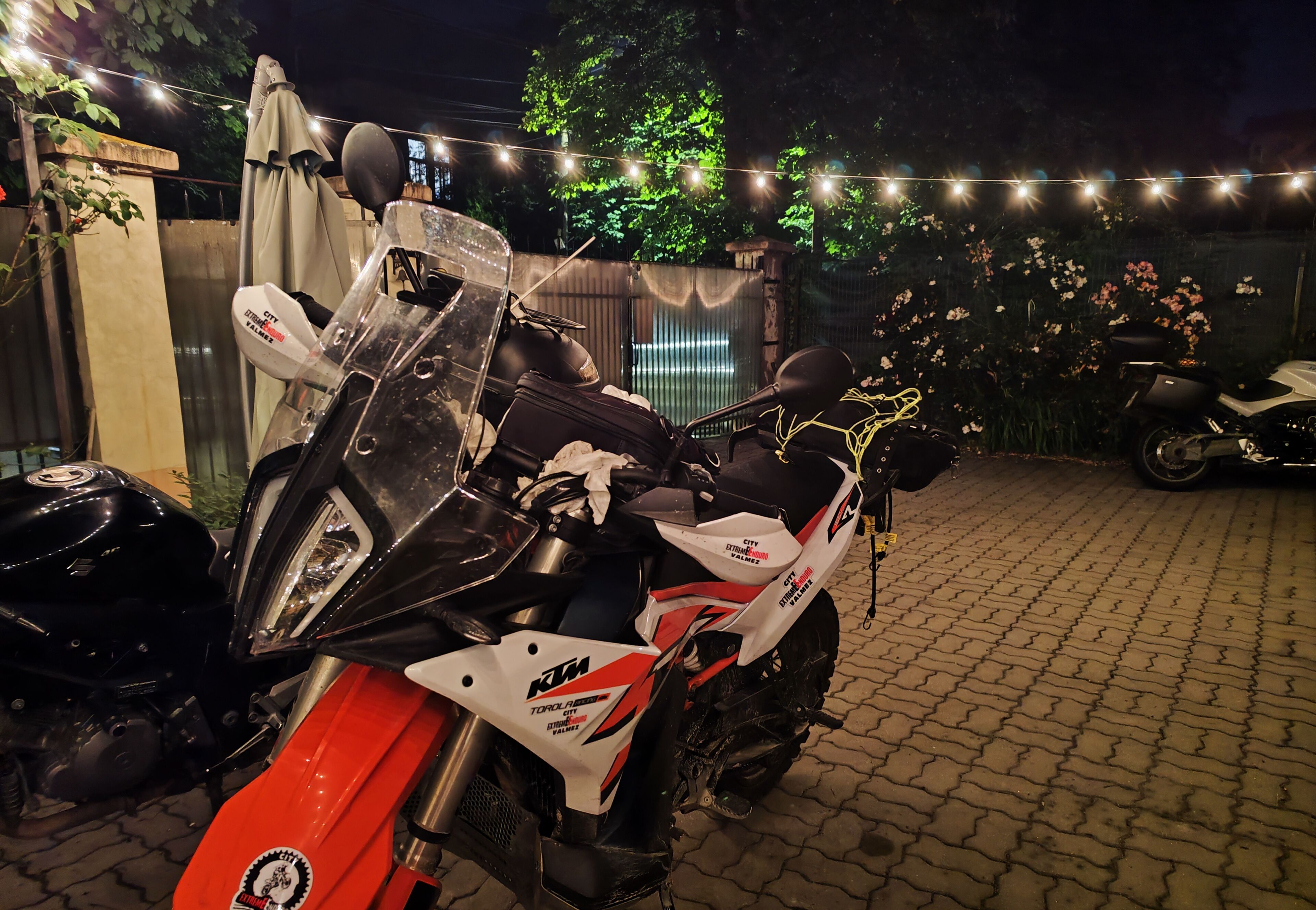 A motorcycle parked in a covered outdoor area with string lights and flowers in the background.