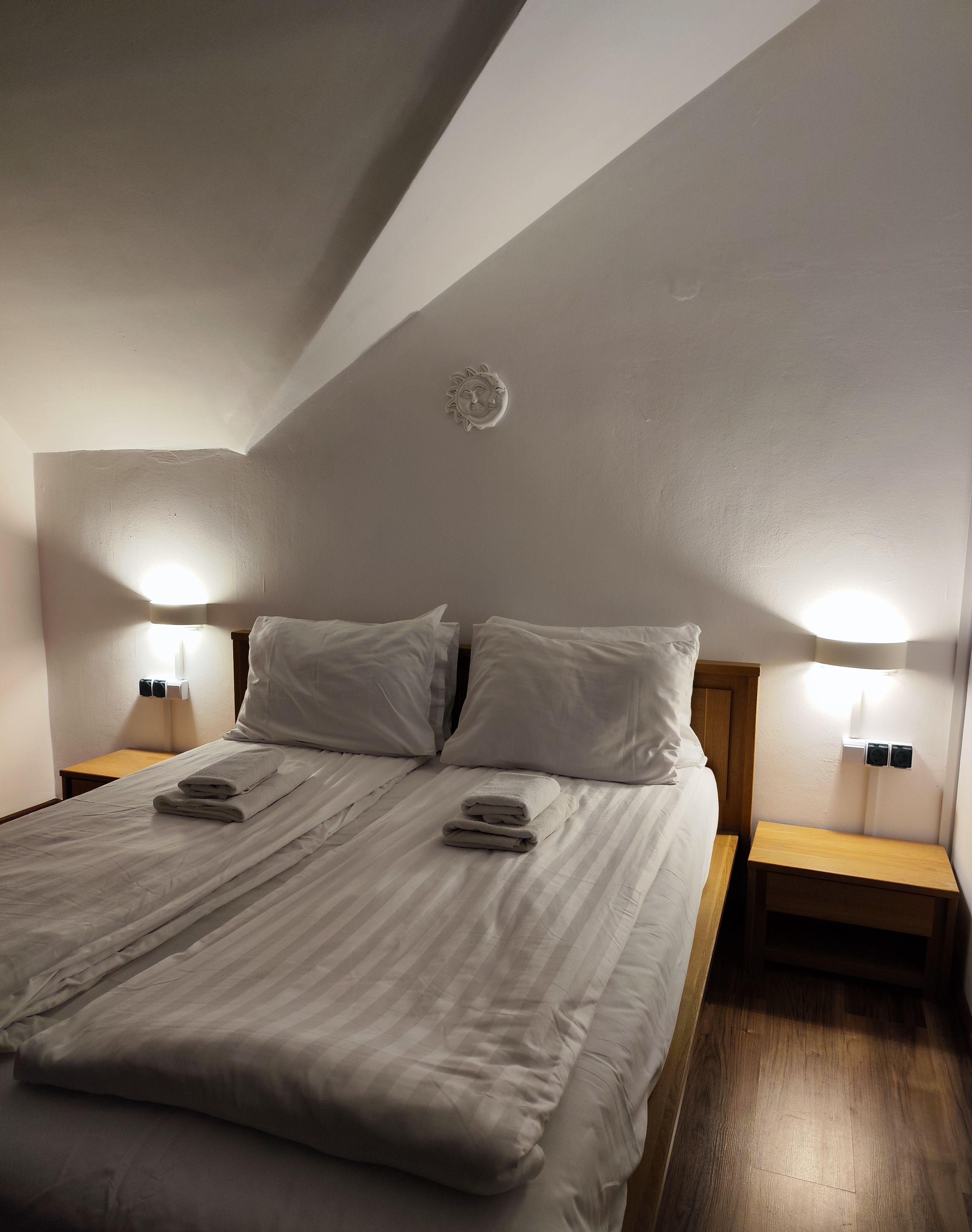 Una cama bien tendida en una habitación con techo inclinado, dos lámparas de noche y una pequeña mesita de madera.
