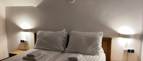 Una cama bien tendida en una habitación con techo inclinado, dos lámparas de noche y una pequeña mesita de madera.