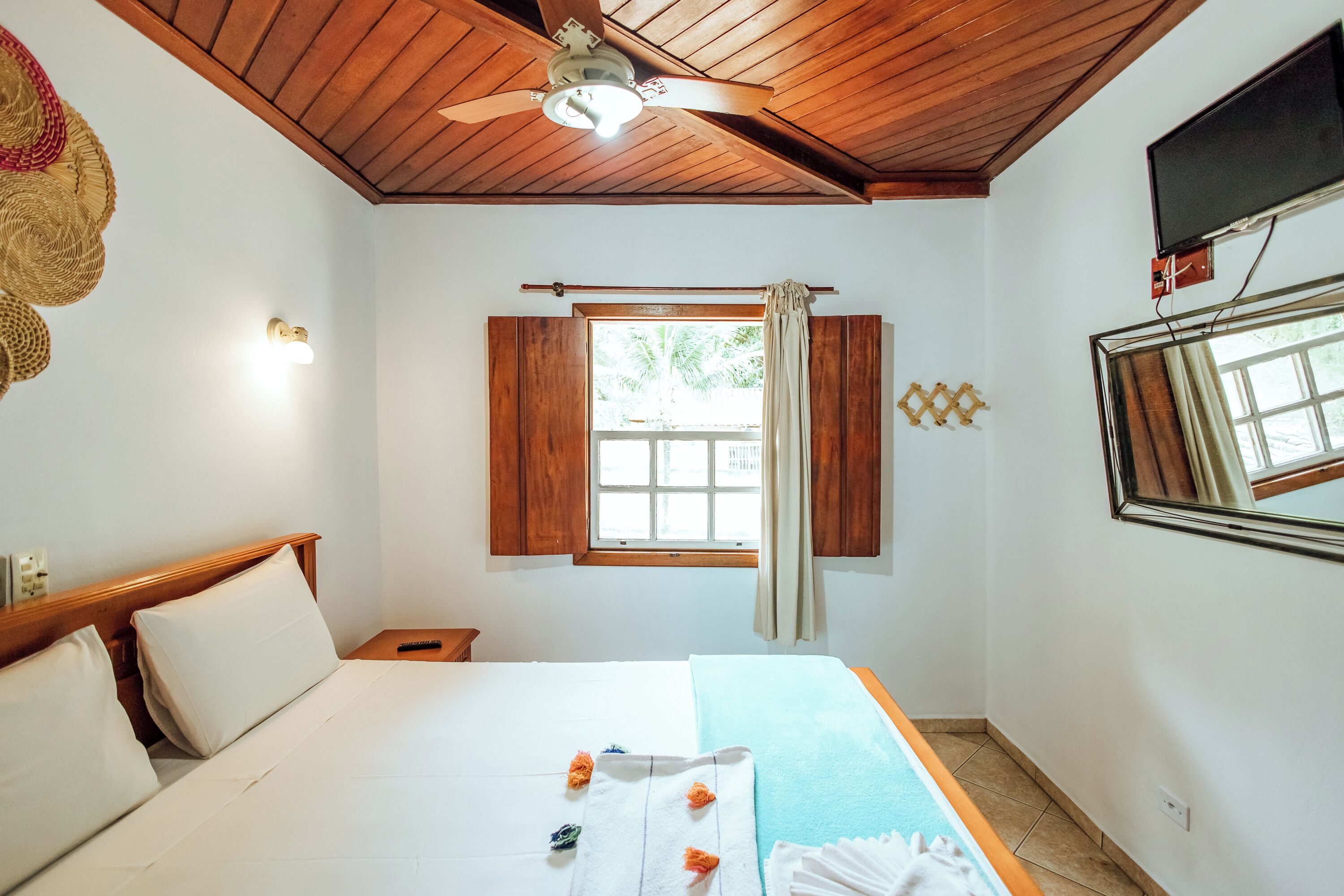 Une chambre d’hôtel avec un plafond en bois, un lit recouvert de draps blancs, une fenêtre avec des rideaux, un téléviseur à écran plat fixé au mur et une petite table de chevet.