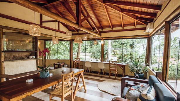 A spacious dining area with a wooden table and chairs, a sofa with cushions, and large windows offering a view of greenery. - Santa Smart Hotel (Ilhabela)