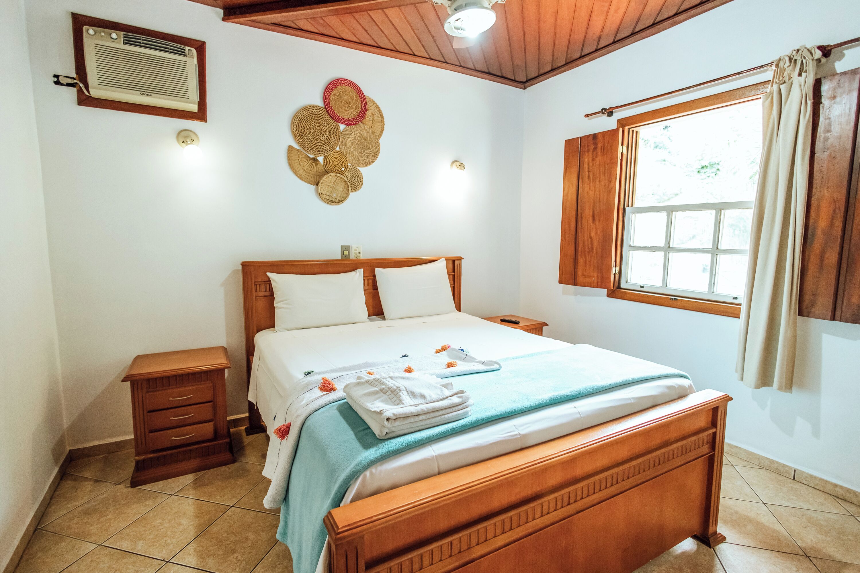 Une chambre avec un lit en bois, une table de chevet, un climatiseur fixé au mur, une fenêtre avec des rideaux et une décoration murale composée de paniers tressés.