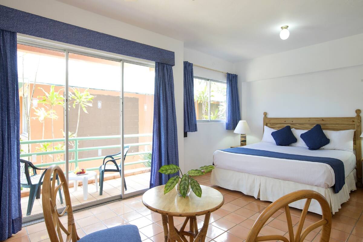 A hotel room with a bed, a table, chairs, and a view of the outside.