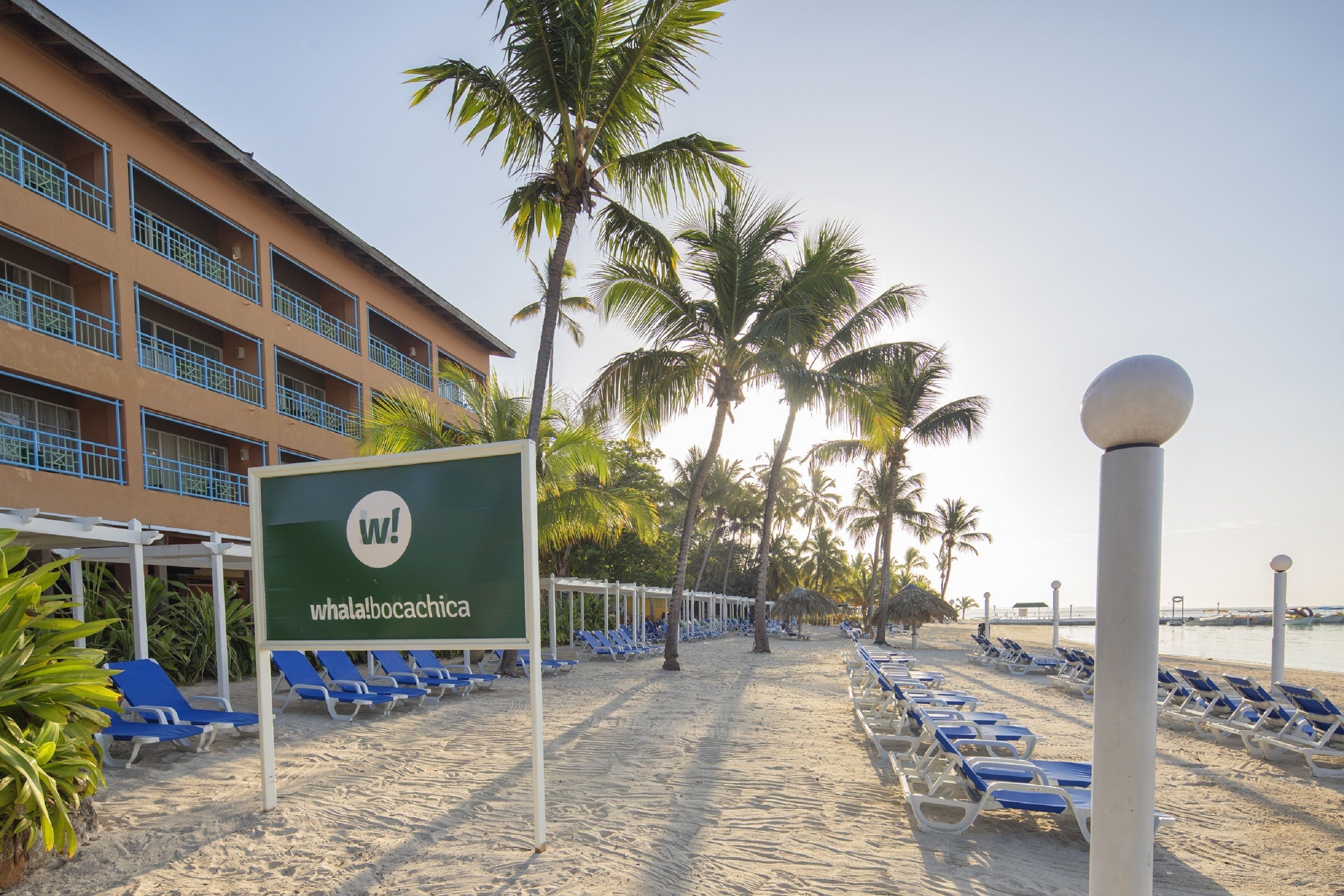 A beachside hotel with a sign reading 'whala!bocachica', blue lounge chairs, palm trees, and a clear sky.