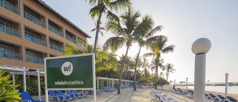 A beachside hotel with a sign reading 'whala!bocachica', blue lounge chairs, palm trees, and a clear sky.