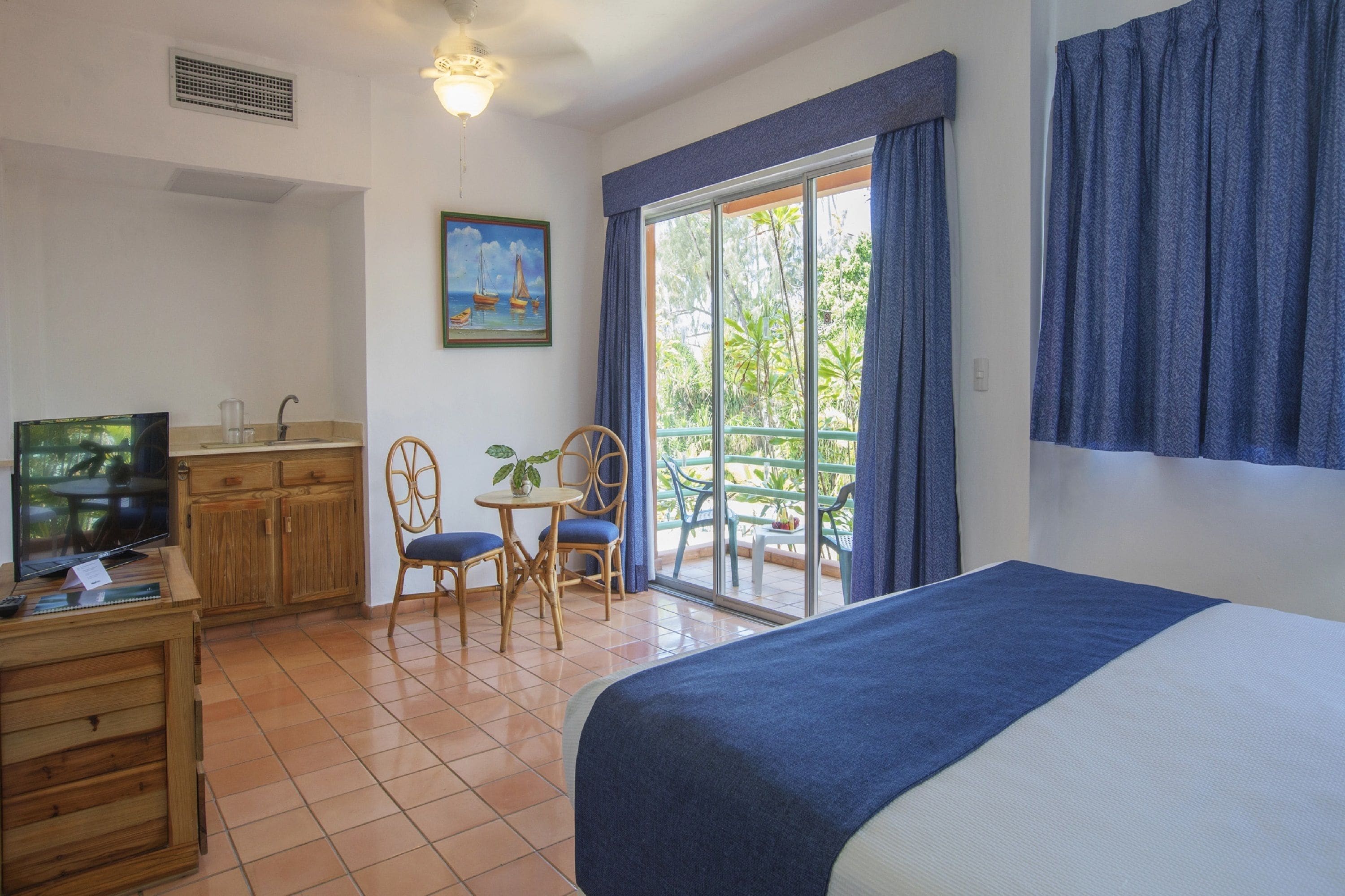 Quarto de hotel com cama, escrivaninha com TV, pequena mesa com cadeiras, janela com vista para o exterior e um quadro na parede.