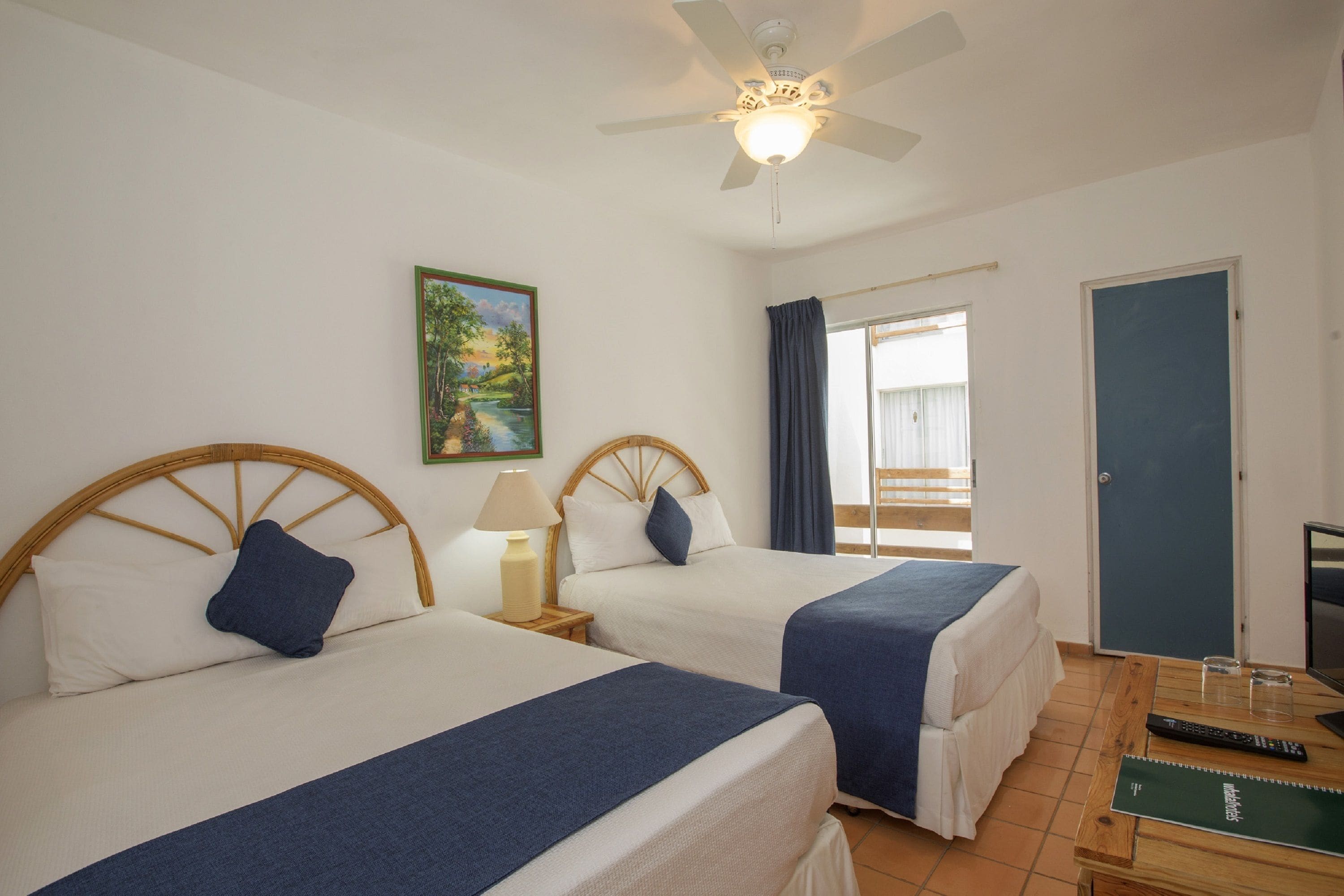 A hotel room with two beds, a ceiling fan, a painting, a window with curtains, and a desk with a computer and a notebook.