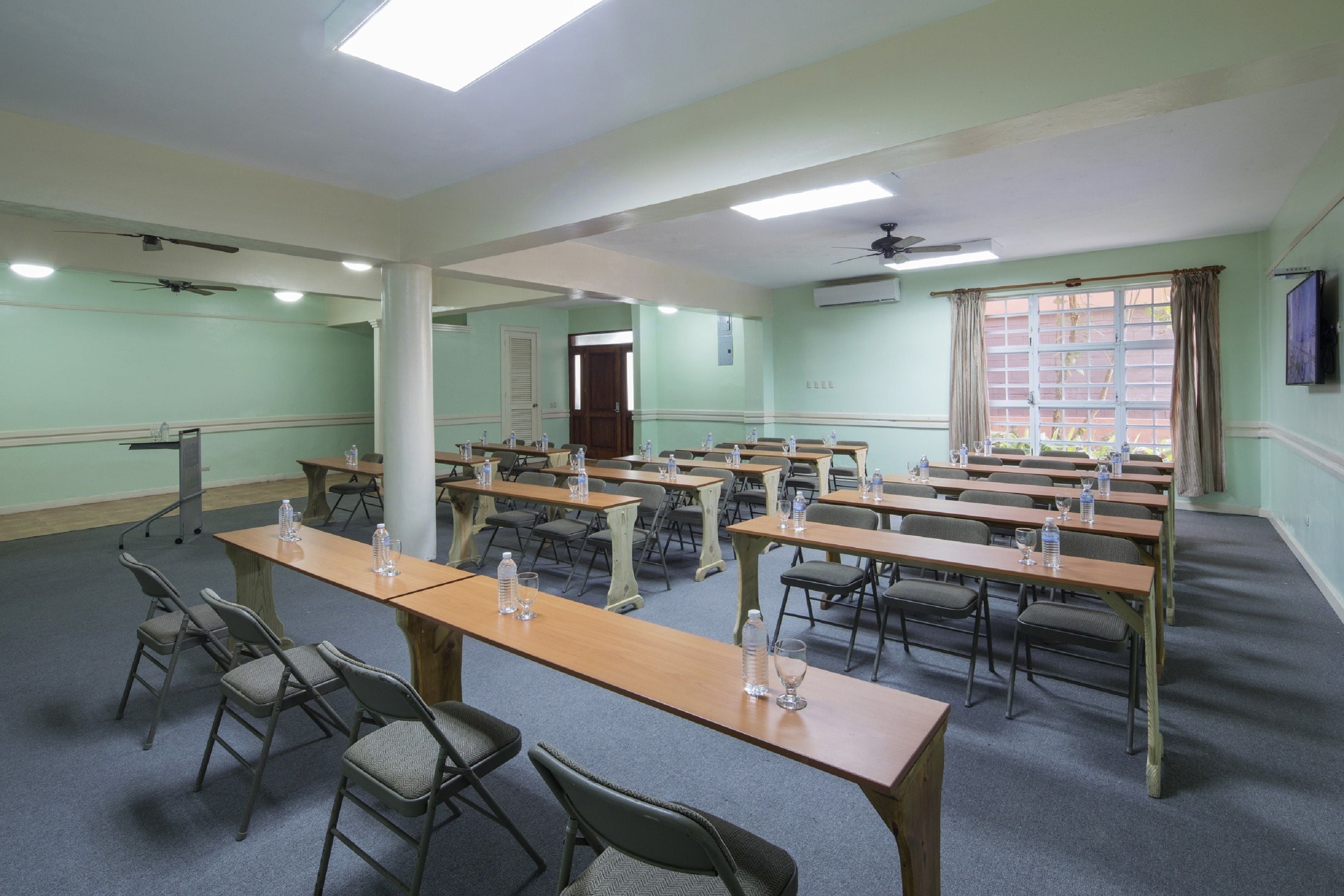 Une salle de conférence équipée de plusieurs tables et chaises, d’un ventilateur de plafond et d’une grande fenêtre avec des rideaux.
