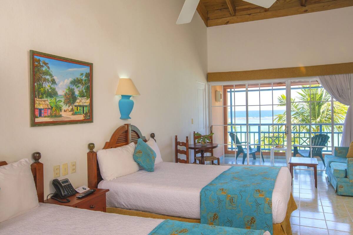 A hotel room with two beds, a painting of a tropical scene, a phone on the nightstand, and a balcony with chairs and a table.