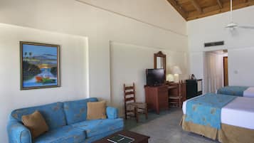 A hotel room with a blue sofa, a desk with a television, a bed with a patterned bedspread, and a painting of a coastal scene.