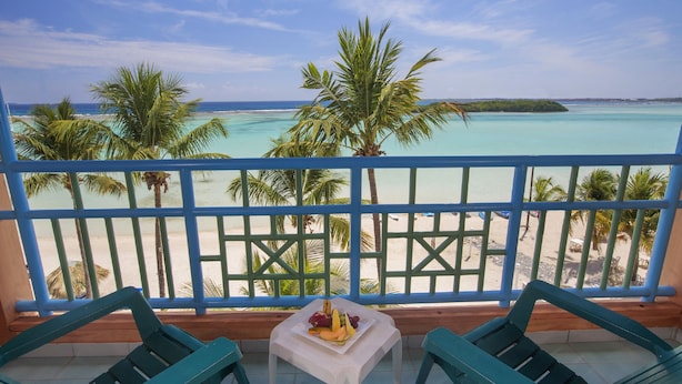 A balcony with a view of a beach and turquoise waters, featuring two green chairs and a small table with fruit.