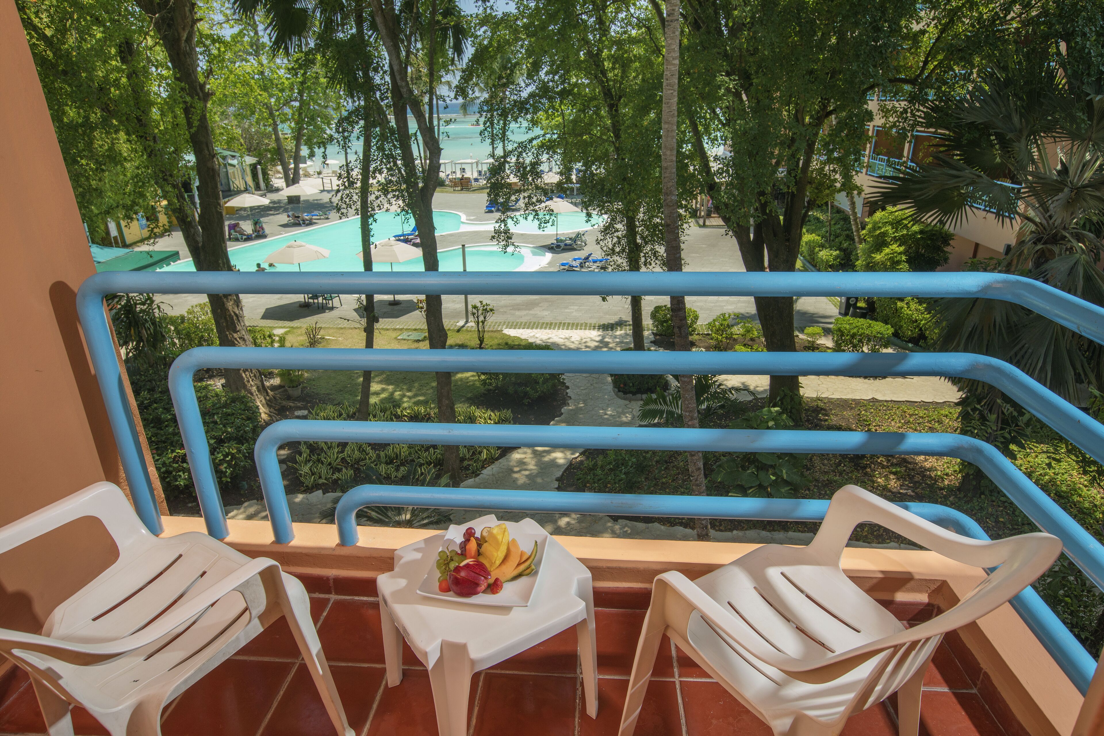 Un balcón con dos sillas de plástico blancas, una mesita con fruta, y vista a una piscina y palmeras.