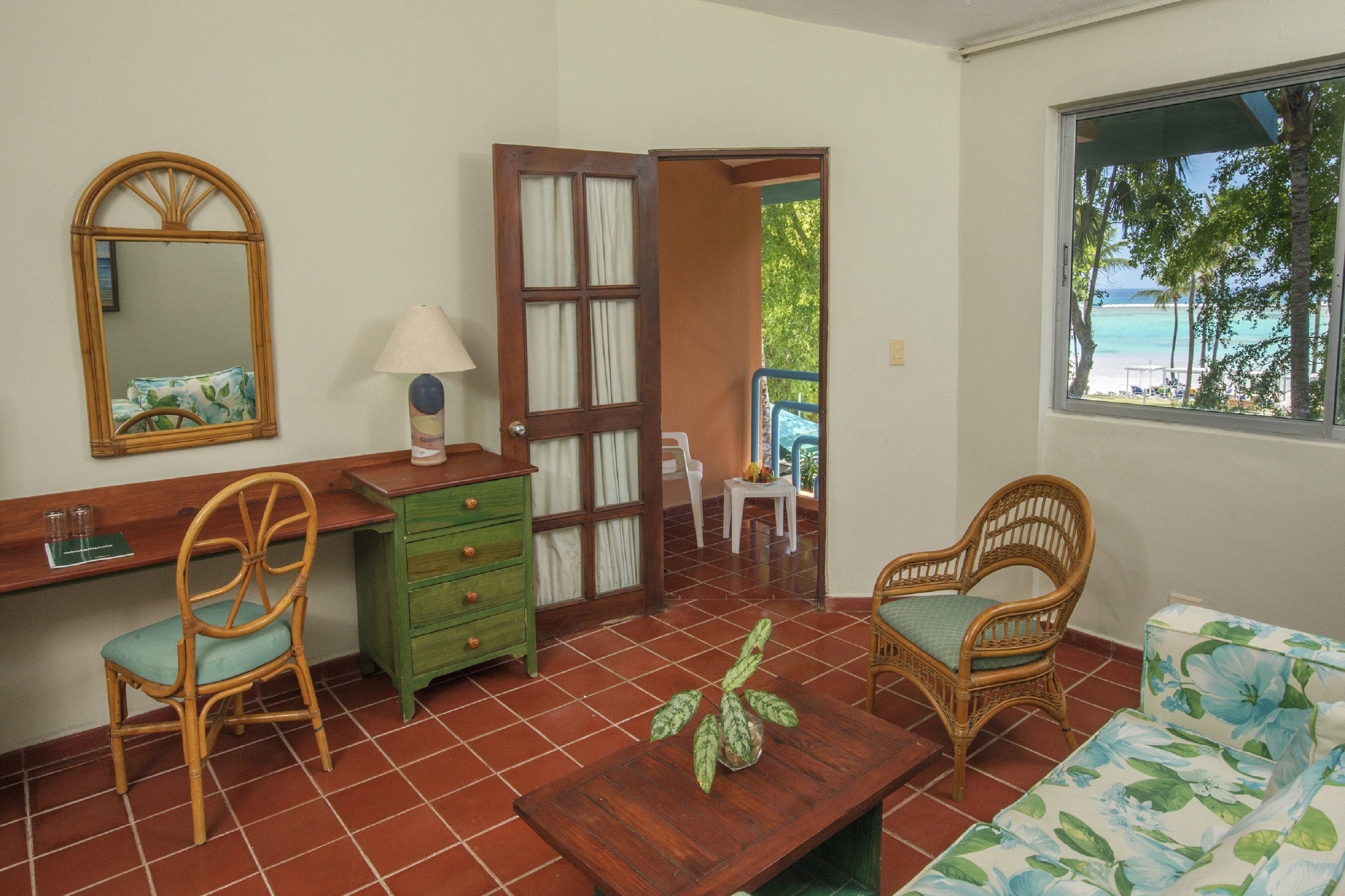 A room with a wooden desk, two wicker chairs, a green dresser, a wooden table with a plant, and a window with a view of the beach.