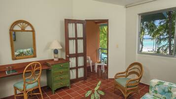 A room with a wooden desk, two wicker chairs, a green dresser, a wooden table with a plant, and a window with a view of the beach.