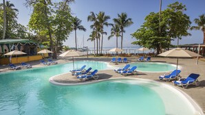 A pool area with blue lounge chairs and umbrellas, surrounded by palm trees and a clear sky.
