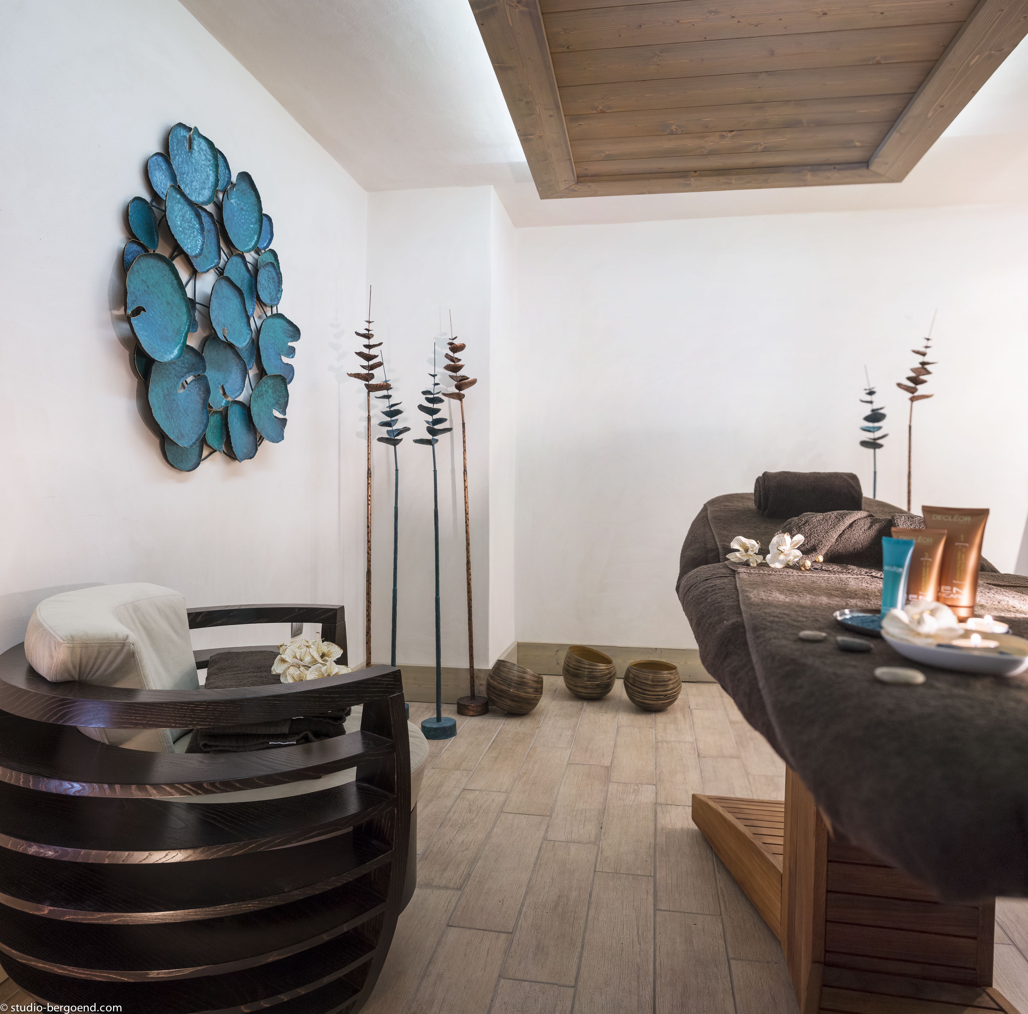A modern spa room with a massage table, a wooden chair, and decorative elements like a blue wall art piece and tall, slender vases.