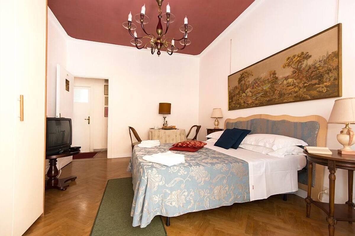A bedroom with a bed, a chandelier, a painting, a television, and a table.