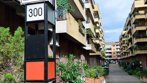 A narrow urban street with apartment buildings on either side, a street sign with the number 300, and potted plants along the sidewalk. - Minerva B&B (Rome)